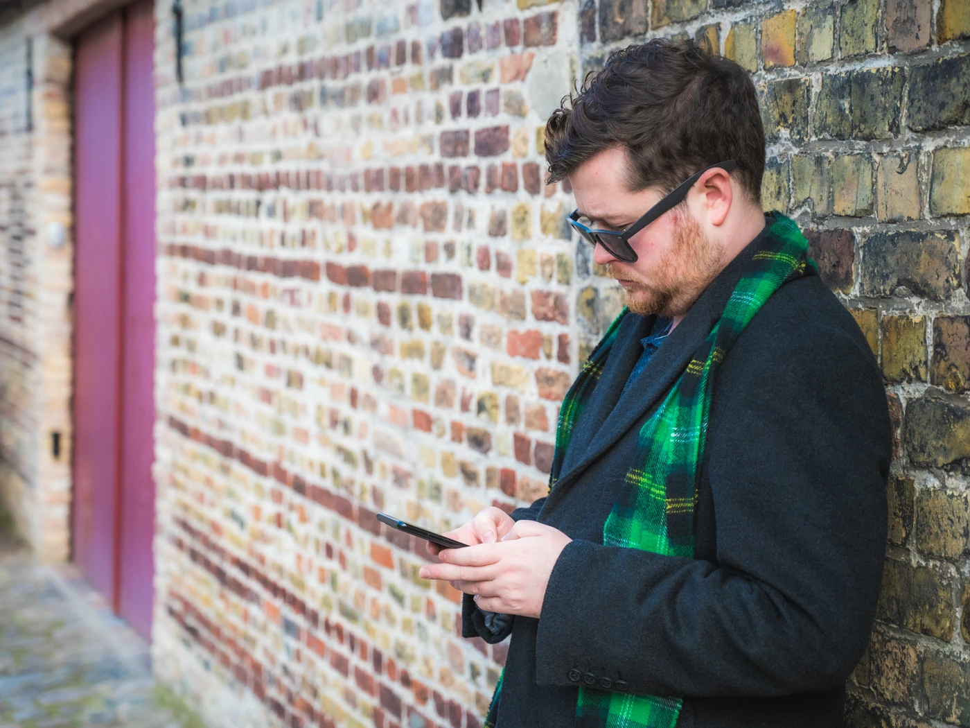 A man with sunglasses, a green scarf and an overcoat leaning against a wall using his phone