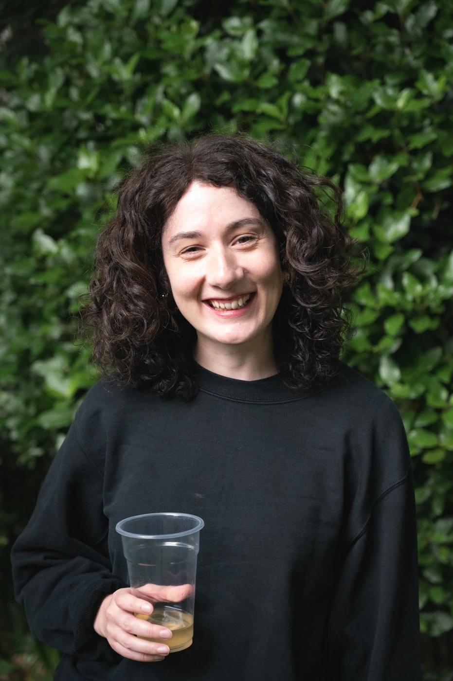 Woman with dark curly hair holding a drink in a plastic cup smiling