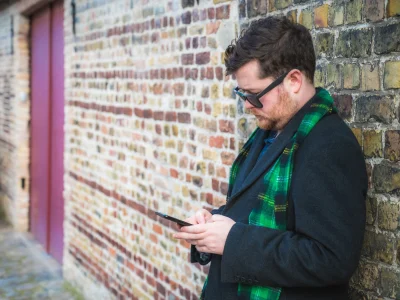 A man with sunglasses, a green scarf and an overcoat leaning against a wall using his phone